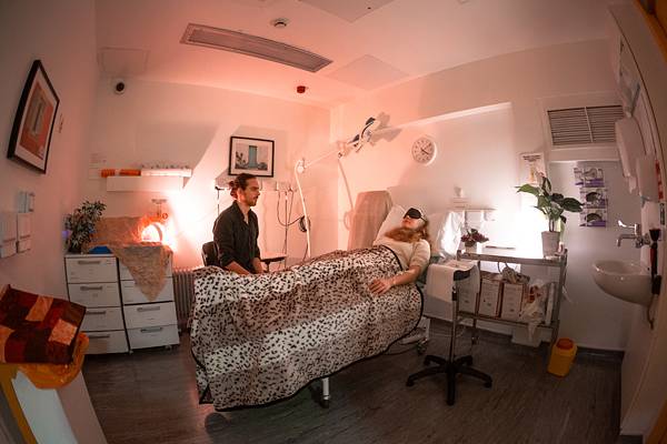 Staged re-enactment of a research session: a person lies on a clinical bed wearing eyeshades under a blanket, accompanied by a researcher seated alongside, in a softly lit room with plants.