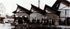 Old sepia photograph of a factory yard with gabled roofs, large spherical vessels, barrels and workers.