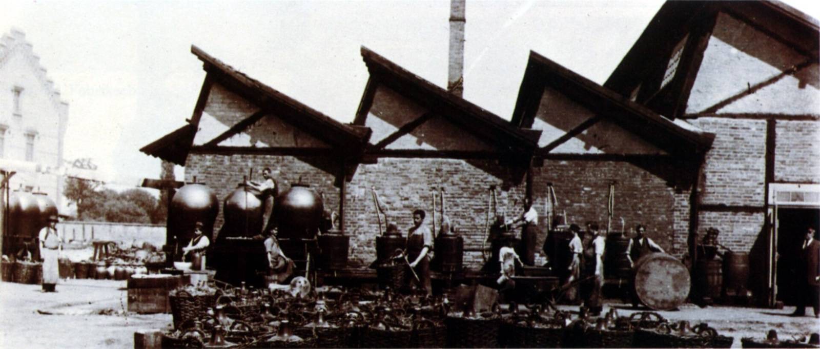 Old sepia photograph of a factory yard with gabled roofs, large spherical vessels, barrels and workers.