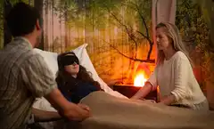 Staged re-enactment of a research session: a person lies on a bed wearing eyeshades under a blanket, accompanied by two therapists sitting beside them, in a softly lit room with a forest mural on the wall.