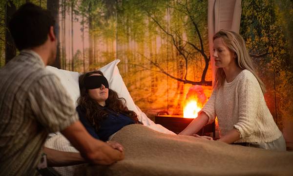Staged re-enactment of a research session: a person lies on a bed wearing eyeshades under a blanket, accompanied by two therapists sitting beside them, in a softly lit room with a forest mural on the wall.